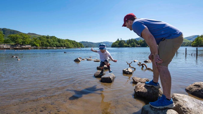 A father and young son standing on stones along the shore of Derwent Water at Crow Park on a sunny day, with a view of the water and land beyond